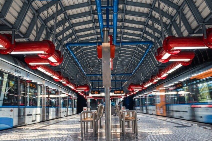 A train station with a covered platform, overhead train tracks, and a moving train. 2