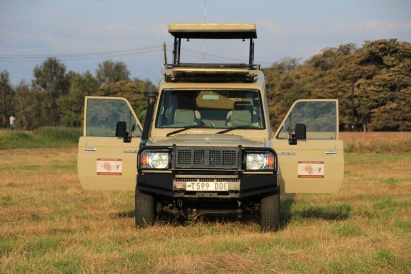 A beige off-road vehicle with an open door, parked in a grassy field with trees in the background. 2