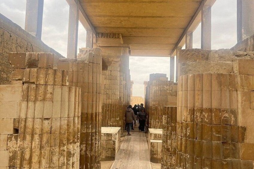 An ancient stone corridor with columns and visitors walking through. 4