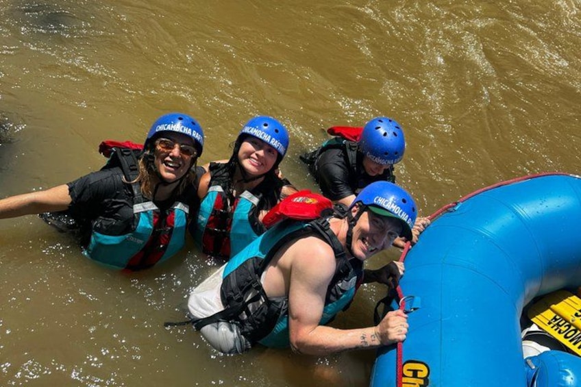 People in water with life vests and helmets, a blue inflatable raft, and a river. 4