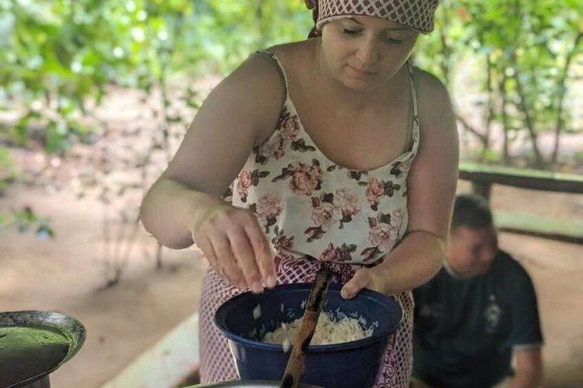 A person preparing food outdoors, wearing a floral dress and a headscarf, with a bowl and a spoon on a table. 1