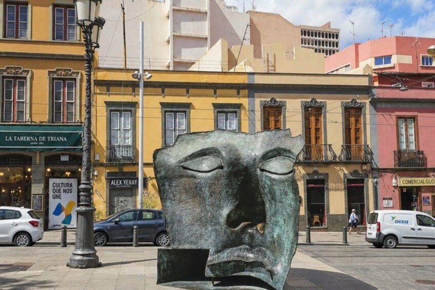 A street scene with a large bronze sculpture of a face, buildings with shops, and parked cars. 4