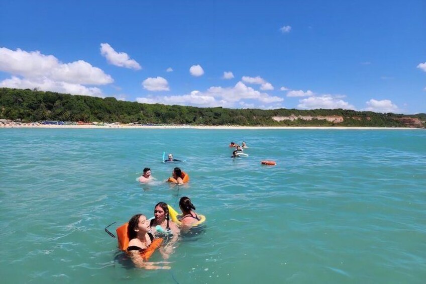 People swimming in clear turquoise water with a beach and greenery in the background. 2