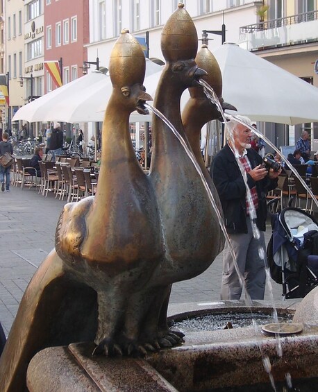 Eine bronzene Skulptur von zwei Seelöwen, aus deren Mündern Wasser fließt, befindet sich in einem öffentlichen Platz mit Cafés und Menschen im Hintergrund. 2