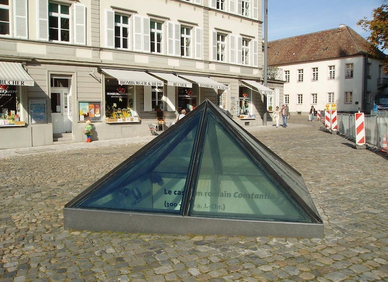 Eine Kopfsteinpflasterstraße mit einer Glaspyramidenstruktur im Vordergrund und Gebäuden mit Beschilderung im Hintergrund. 4