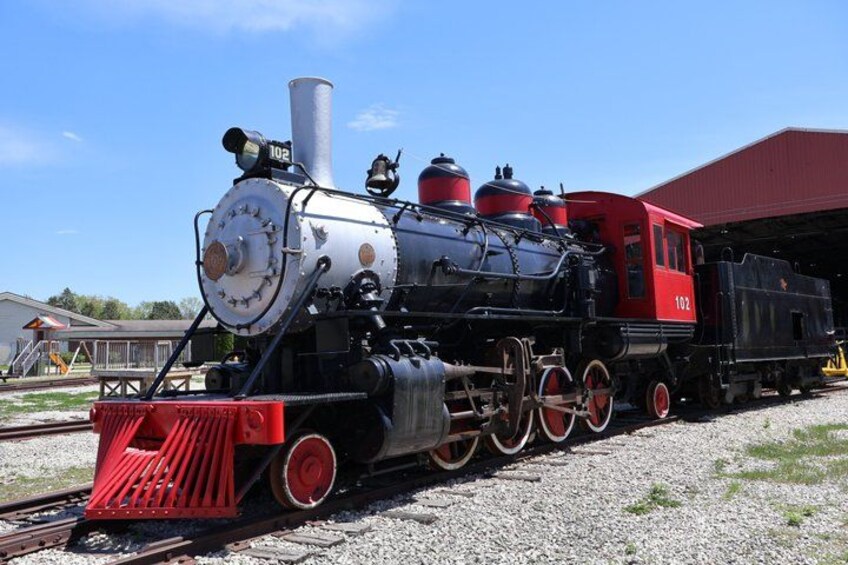 A vintage steam locomotive with the number 102, parked on a railway track with a red building in the background. 5
