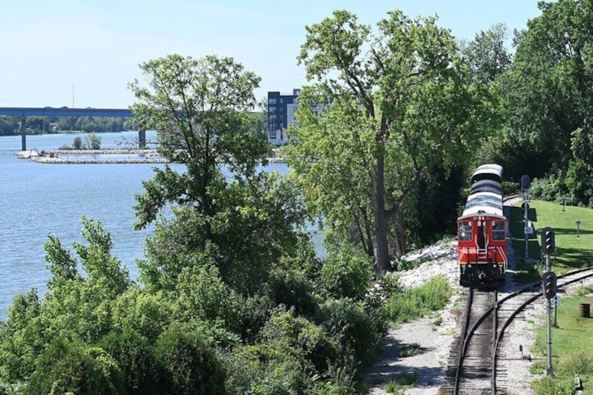 A red train on tracks near a body of water with trees and a bridge in the background. 4