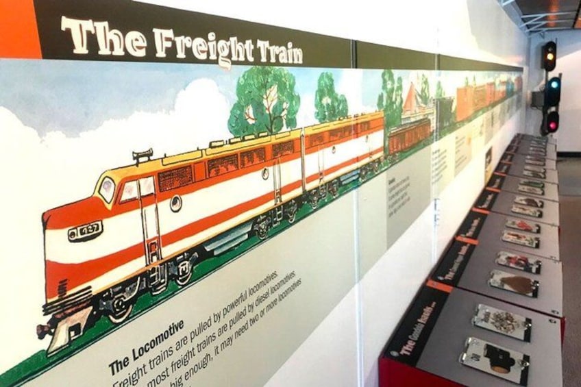 A freight train with a red and white locomotive, a display board with information, and a track with train parts. 3