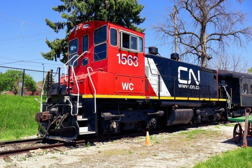 A CN train numbered 1563 on tracks with a red locomotive and black carriages, surrounded by greenery and trees. 2