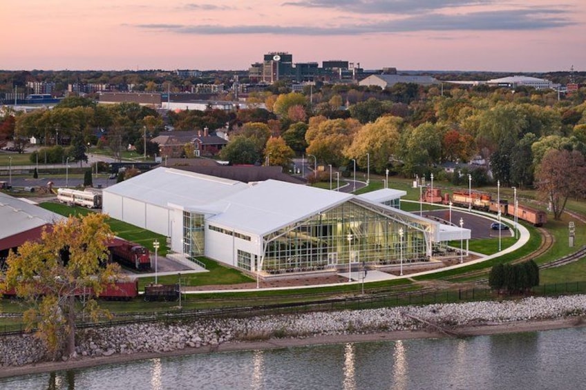 A modern building with a glass facade, surrounded by a park and a body of water, with a cityscape in the background. 1