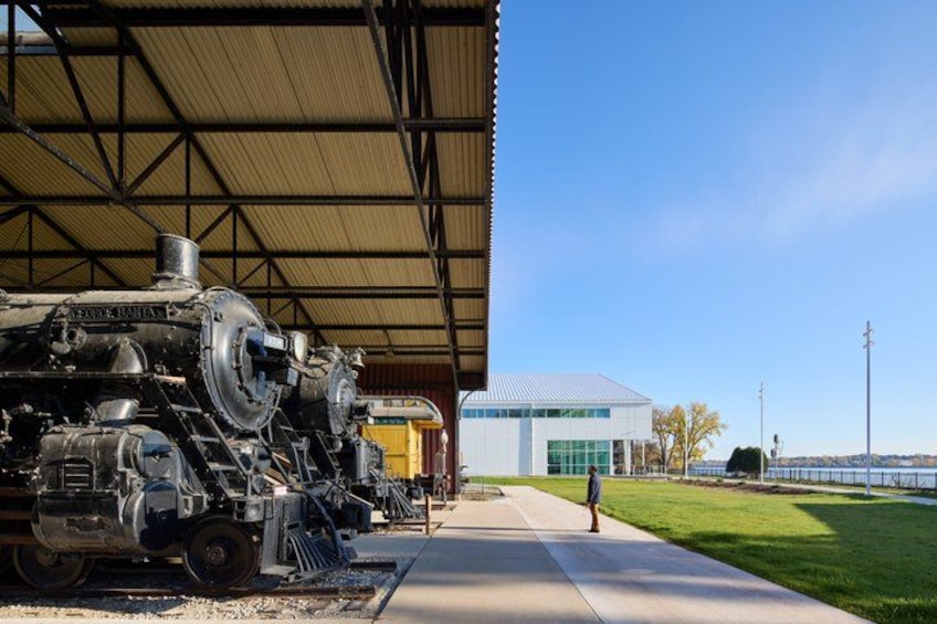 A steam locomotive under a metal roof, a modern building, and a grassy area with a clear sky. 1