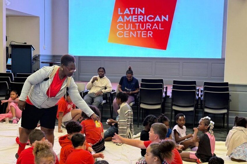 A group of children sitting on the floor in a room with a large screen displaying 'LATIN AMERICAN CULTURAL CENTER'. 1