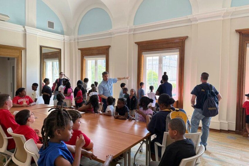 A group of people in a large room with high ceilings and arched windows, some standing and some seated at a long table. 3
