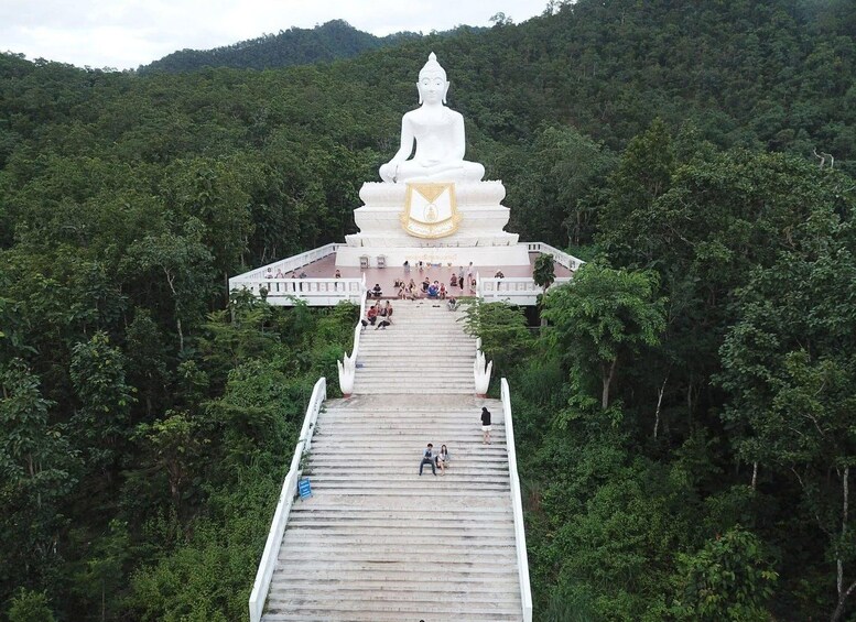 Un grand stupa blanc, surmonté d’un emblème doré, entouré d’une forêt verdoyante et luxuriante, et un long escalier menant à son sommet. 1