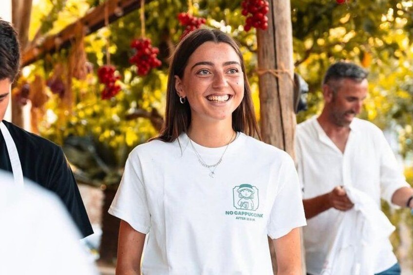 A person wearing a white t-shirt with a logo and text, standing outdoors with trees and red flowers in the background. 1