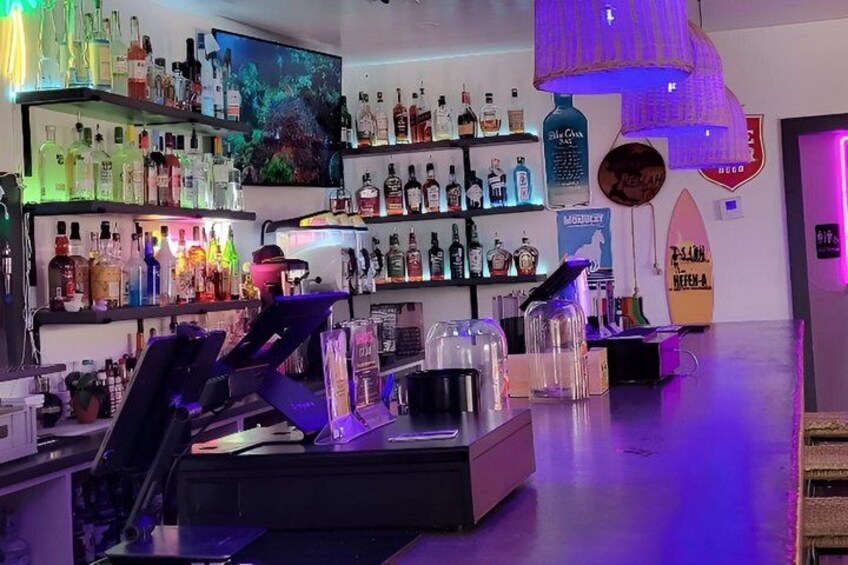 A bar with shelves of liquor bottles, a surfboard decoration, and a neon sign. 5