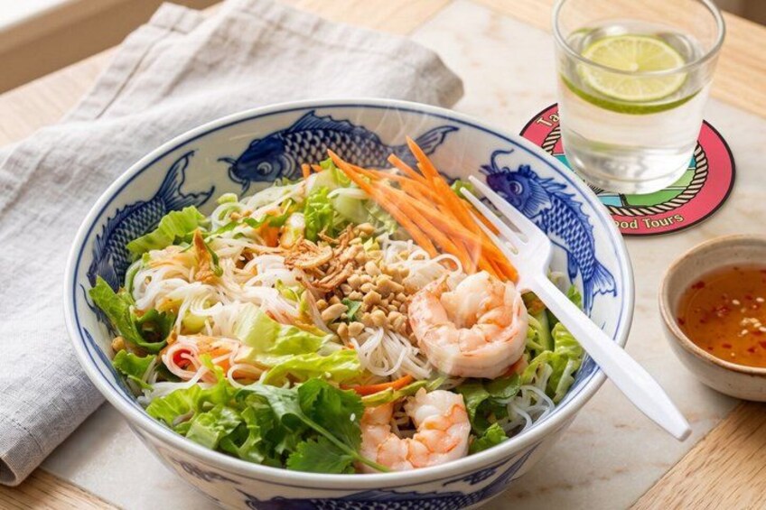 A bowl of noodle salad with shrimp, carrots, and peanuts, accompanied by a glass of lime water and a small bowl of sauce. 5