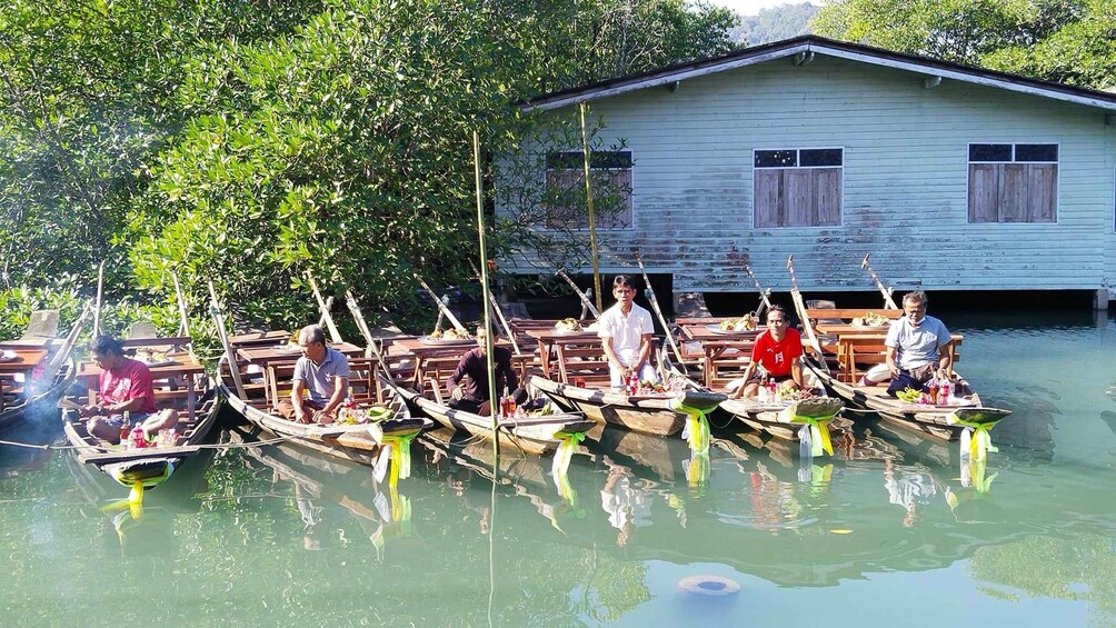 Several boats docked at a wooden pier with people on them, surrounded by lush greenery and a house in the background. 3