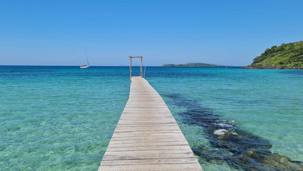 A wooden pier extending into clear turquoise waters with a sailboat in the distance and a lush green island on the right. 3