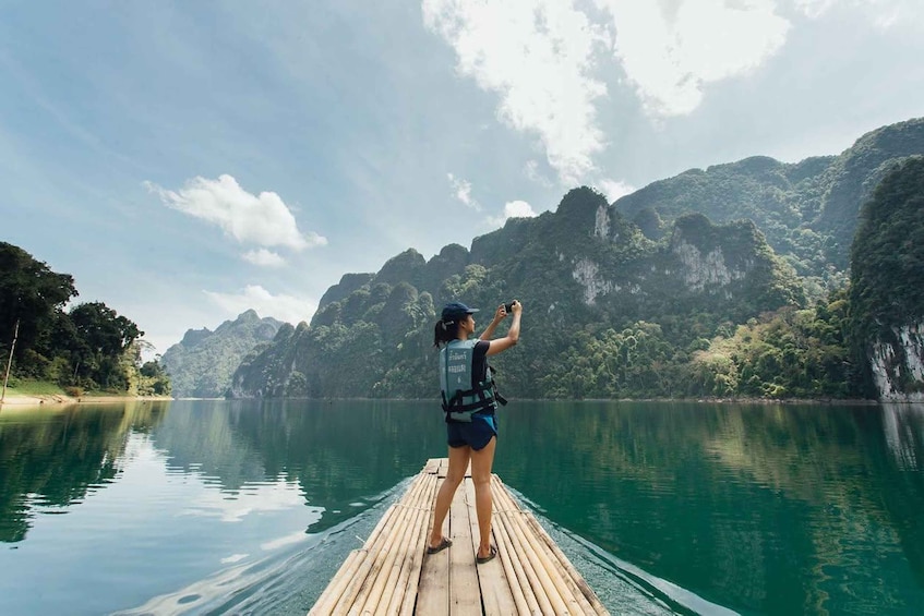 A person standing on a wooden dock extending over a calm lake, surrounded by lush green mountains and clear skies. 2