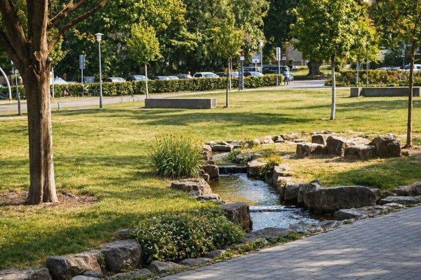 A park with a small stream, rocks, grass, trees, and a paved path. 4