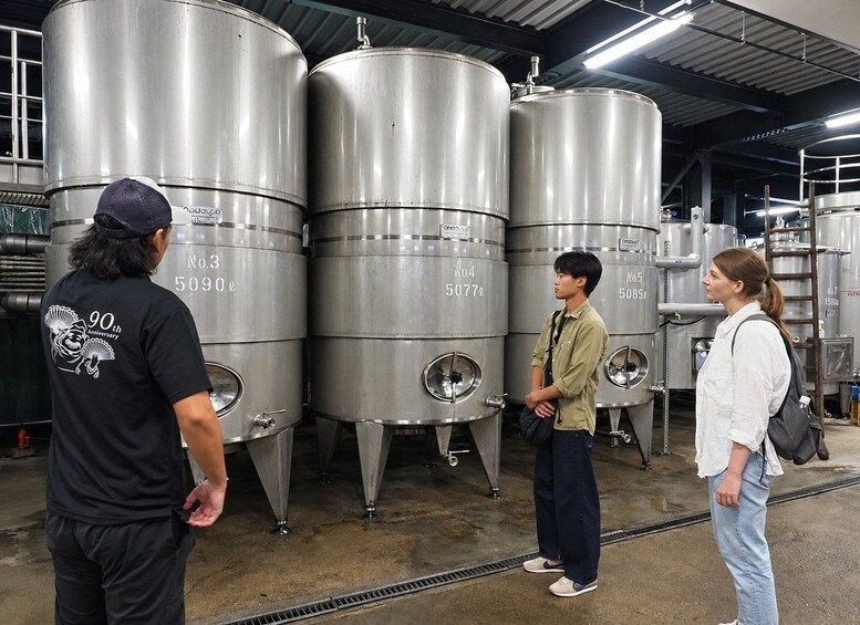Three people standing in a brewery with stainless steel fermentation tanks. 4