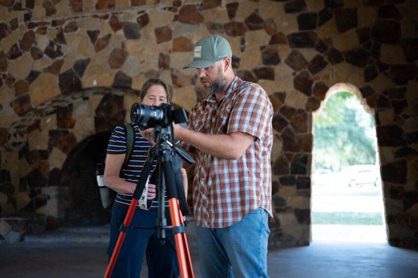A man with a camera on a tripod, a woman with a backpack, and a stone archway in the background. 1