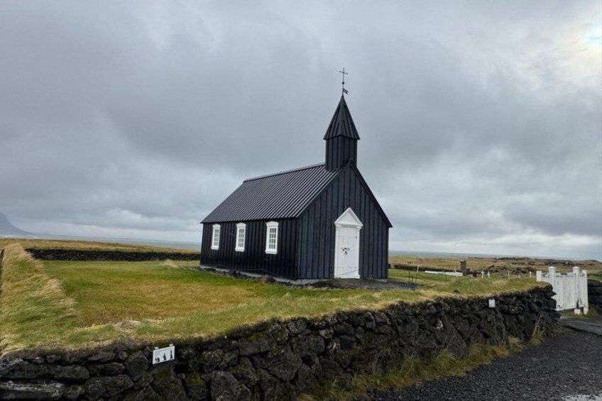 A black church with a white door and a cross on top, surrounded by a stone wall and grassy fields. 3