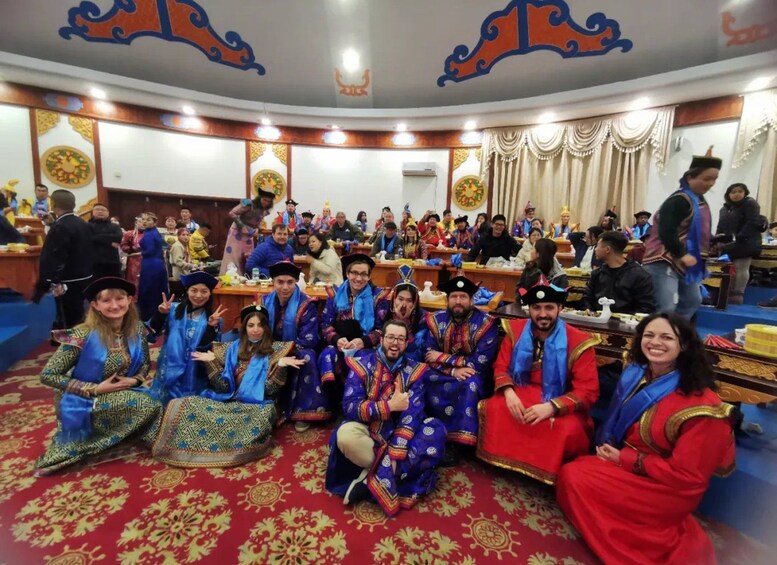 Un groupe de personnes, vêtues de costumes traditionnels, est assis et se tient debout dans un hall décoré, orné de motifs complexes au plafond et de tapis à motifs. 3