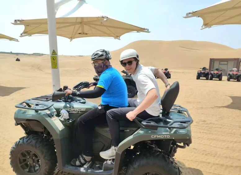Deux personnes se trouvent à bord d’un véhicule tout-terrain vert dans un désert, avec des dunes de sable et d’autres véhicules tout-terrain en arrière-plan. 2