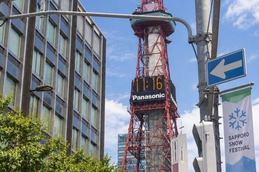 A tall red tower with a digital clock displaying '11:16' and the word 'Panasonic' on it. There is a blue directional sign with an arrow pointing left. 3