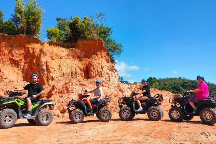 Four people riding atv's on a dirt path with a red dirt hill in the background. 1
