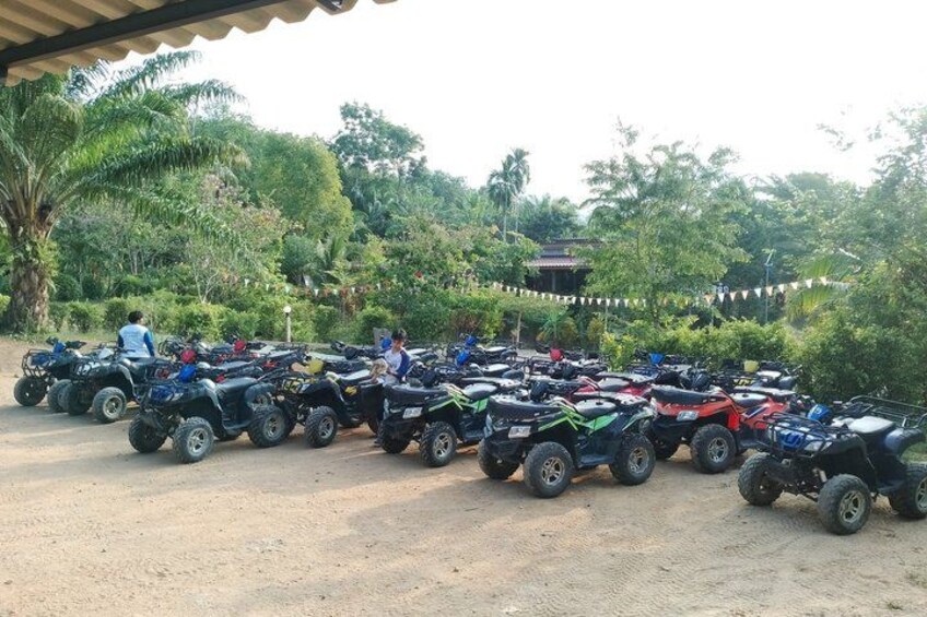 A row of ATVs parked on a dirt path with greenery and a building in the background. 4
