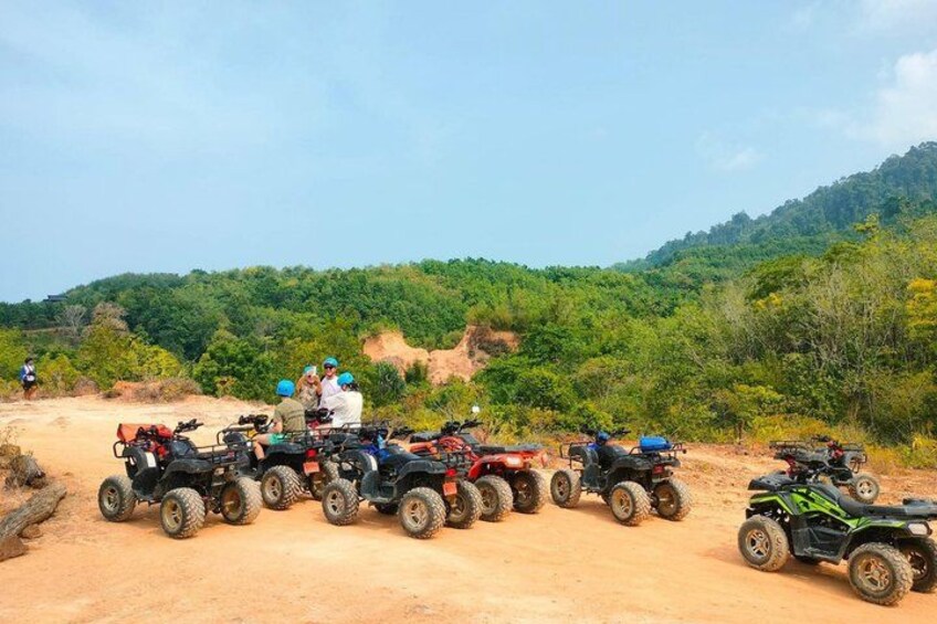A group of off-road vehicles parked on a dirt road with a hilly, forested landscape in the background. 2