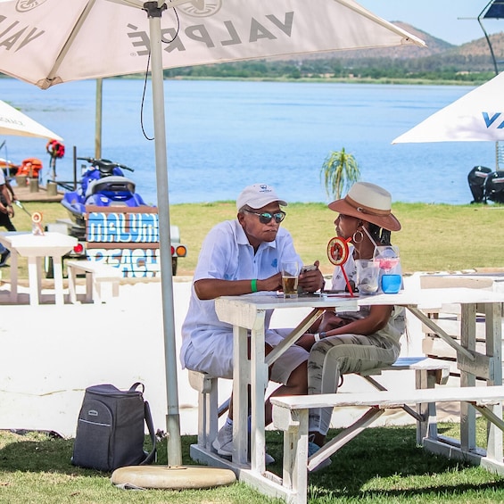 Zwei Personen sitzen an einem Picknicktisch mit Getränken, im Hintergrund ein Strand und in der Nähe ein blaues Fahrzeug. 3
