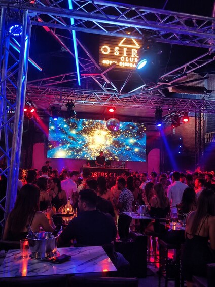 Un club bondé avec une cabine de DJ, des enseignes au néon et un grand écran diffusant une scène cosmique. 2