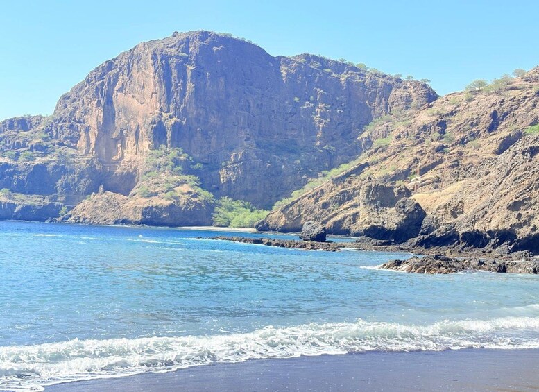 A coastal landscape with a large rocky cliff, clear blue sea, and sandy beach. 3