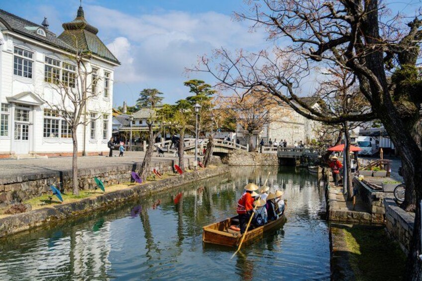 A canal with a boat carrying people, a white building, and a tree with bare branches. 1