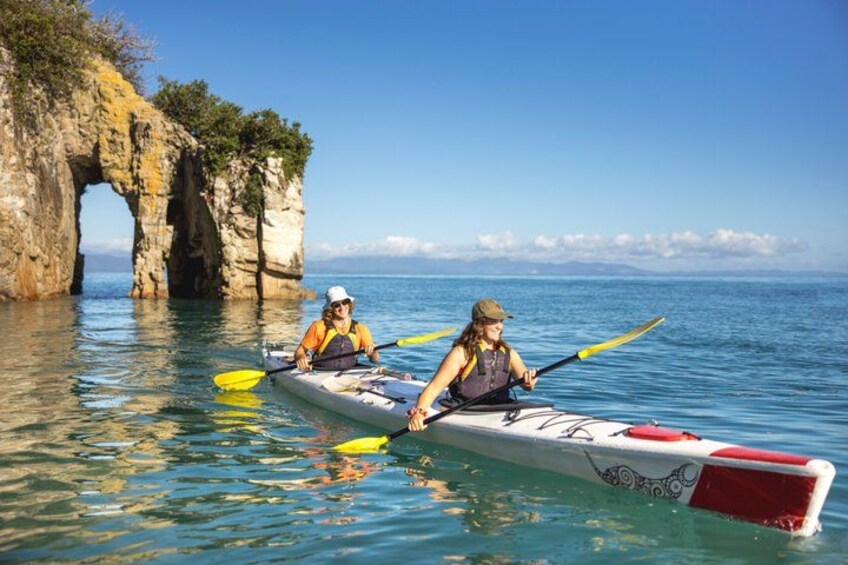 Two people kayaking on a calm sea with a rocky arch formation in the background. 1
