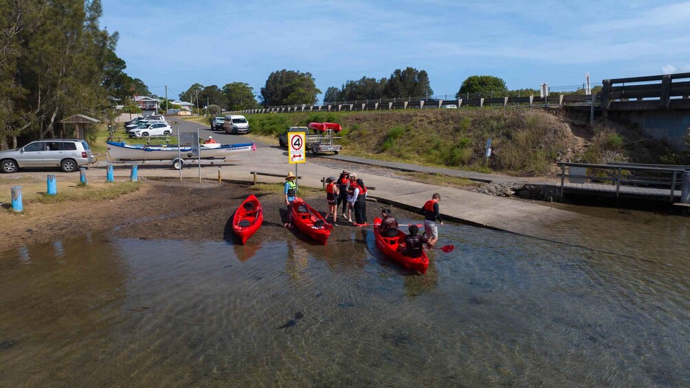 A group of people in red kayaks at a river crossing with a speed limit sign and a bridge in the background. 5