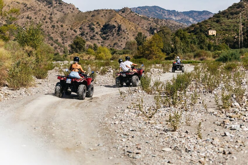 Three people riding ATVs on a rocky dirt road with mountains in the background. 5