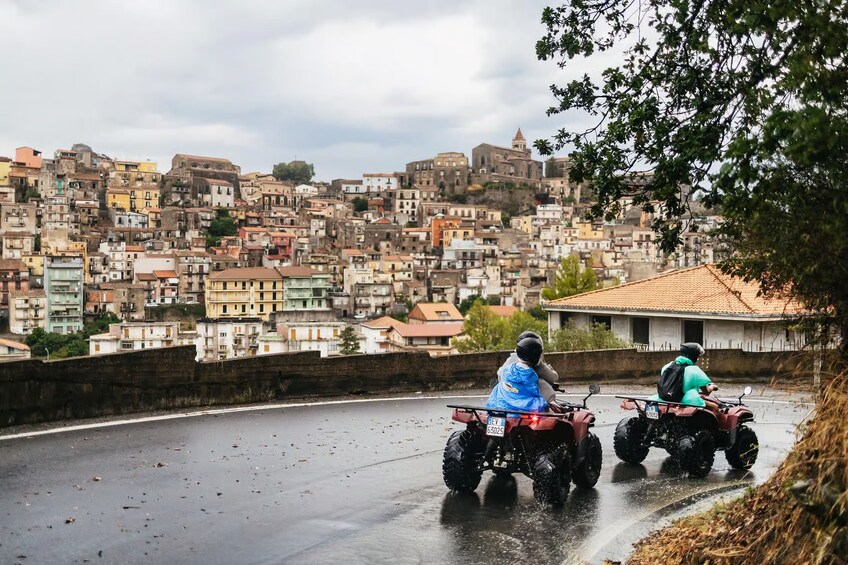 Two riders on quad bikes on a wet road with a densely packed urban landscape in the background. 4