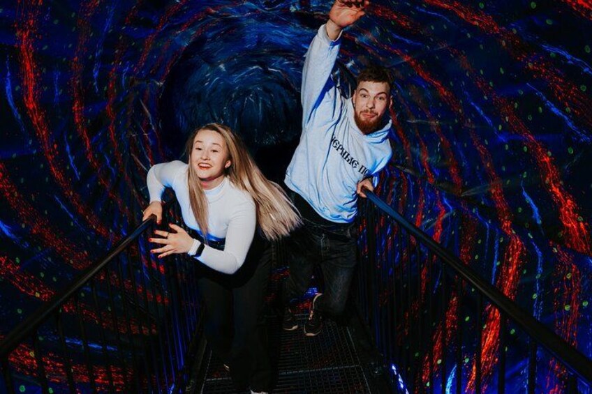 Two people holding onto a railing in a tunnel with a swirling pattern on the walls. 5