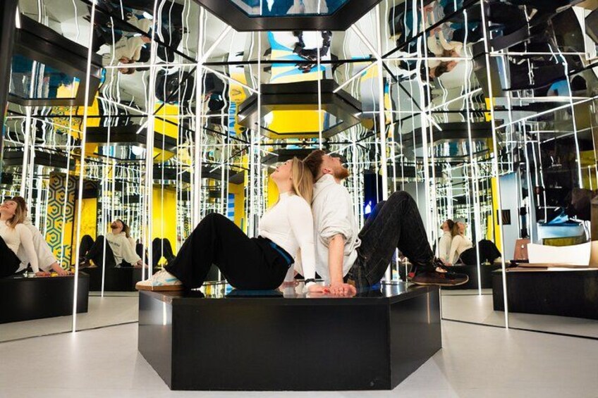 A group of people sitting on a black platform in a modern, geometric interior with reflective surfaces and yellow accents. 1