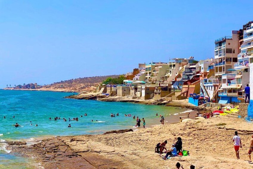 A beach with people swimming and sunbathing, buildings along the shore, and a clear blue sky. 5