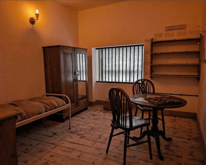 A room with a wooden cabinet, a bed, a table with chairs, and a window with bars. 1