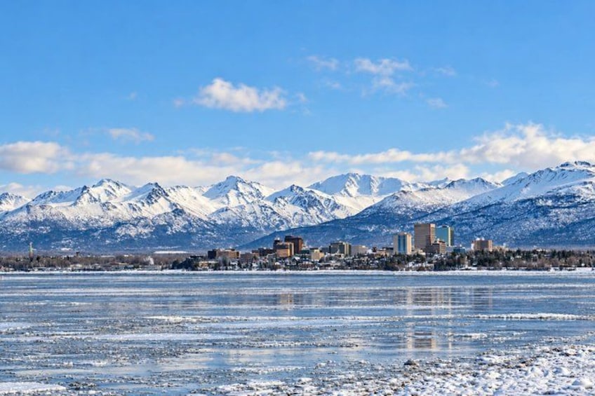A cityscape with snow-covered mountains in the background, a frozen body of water in the foreground, and several buildings. 3