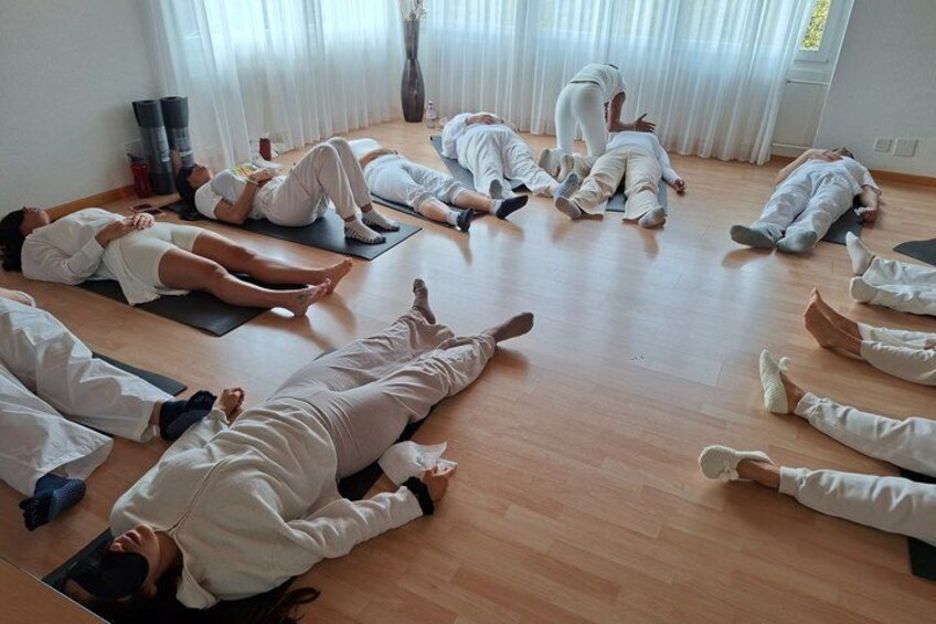 A group of people in white attire lying on mats in a room with wooden flooring and white curtains. 1