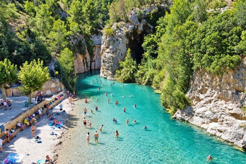 A turquoise river surrounded by rocky cliffs and lush greenery, with people swimming and sunbathing on the banks. 4