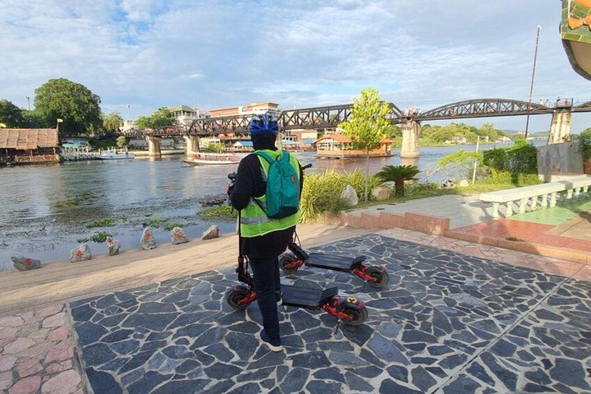 A person on a scooter with a backpack, a river, a bridge, and a stone-paved area. 2